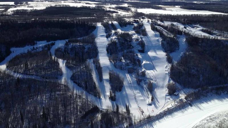 An image of the Nitehawk ski resort in Northern Alberta, Canada, showcasing a picturesque winter sport scene. The resort's chalet sits at the foot of the snow-covered slopes with a skier trailing down.