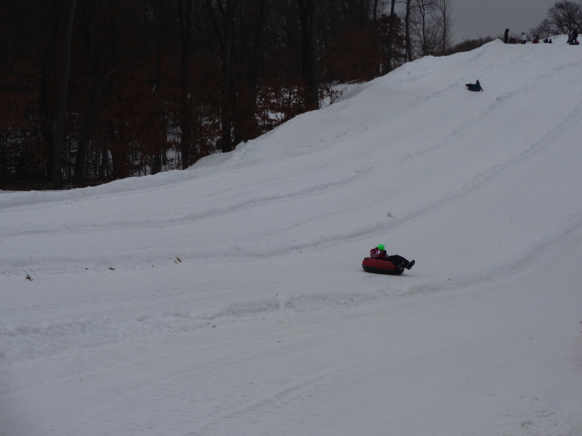 A active winter sports scene at Christmas Mountain, Wisconsin, featuring a snowmobile and a skier in motion. A quaint chalet and ski lift are visible in the background.