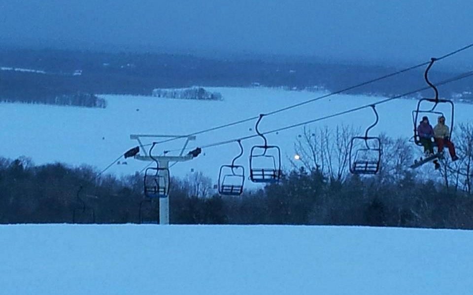 A ski lift operating at Hermon Mountain in Maine USA during winter. There are several skiers enjoying their time including a family and a larger group amidst a vibrant winter sports scene.