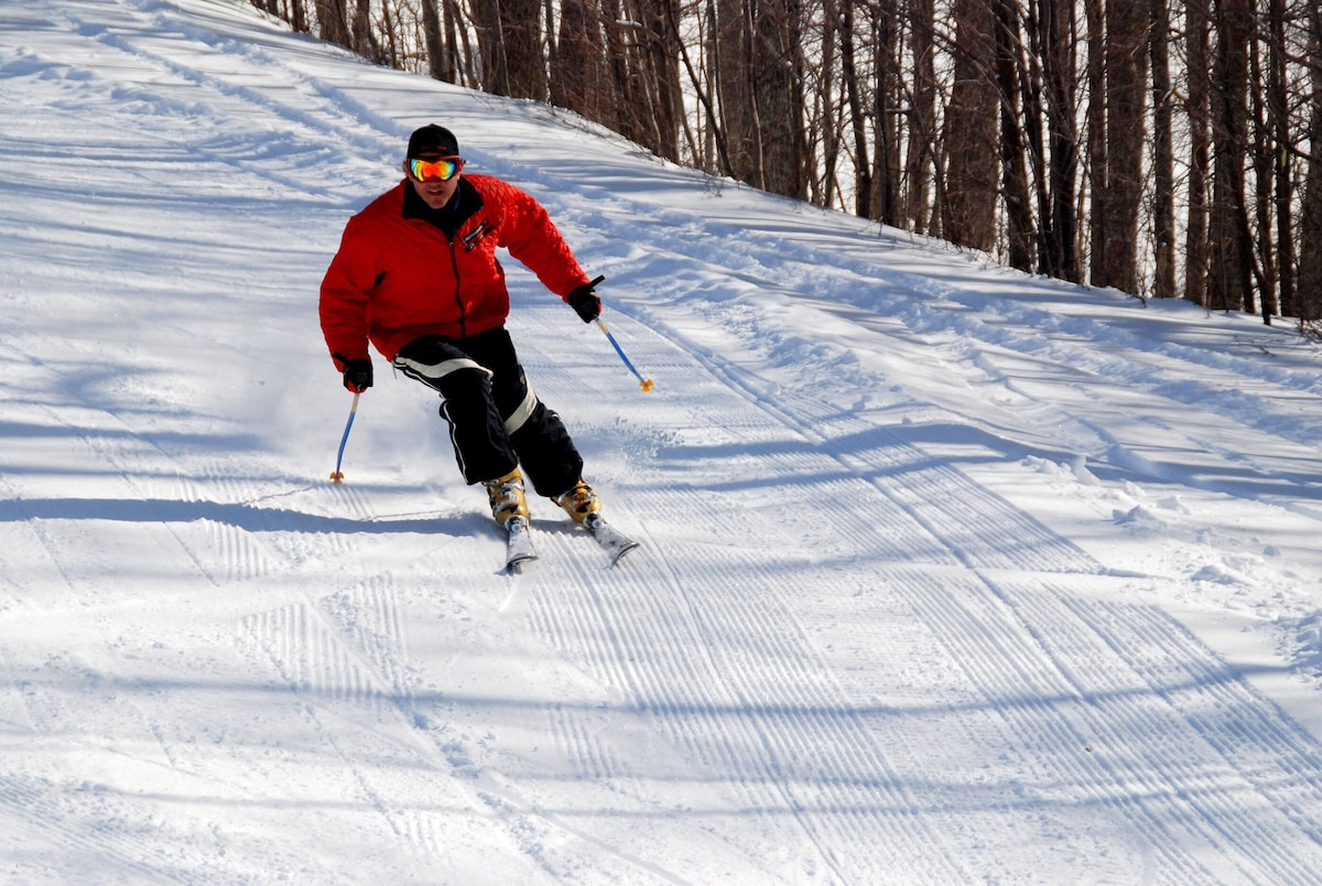A winter sports scene at The Omni Homestead in Virginia with a skier and a snowboarder navigating the snowy slopes. A chalet and a snowmobile are faintly visible in the background.
