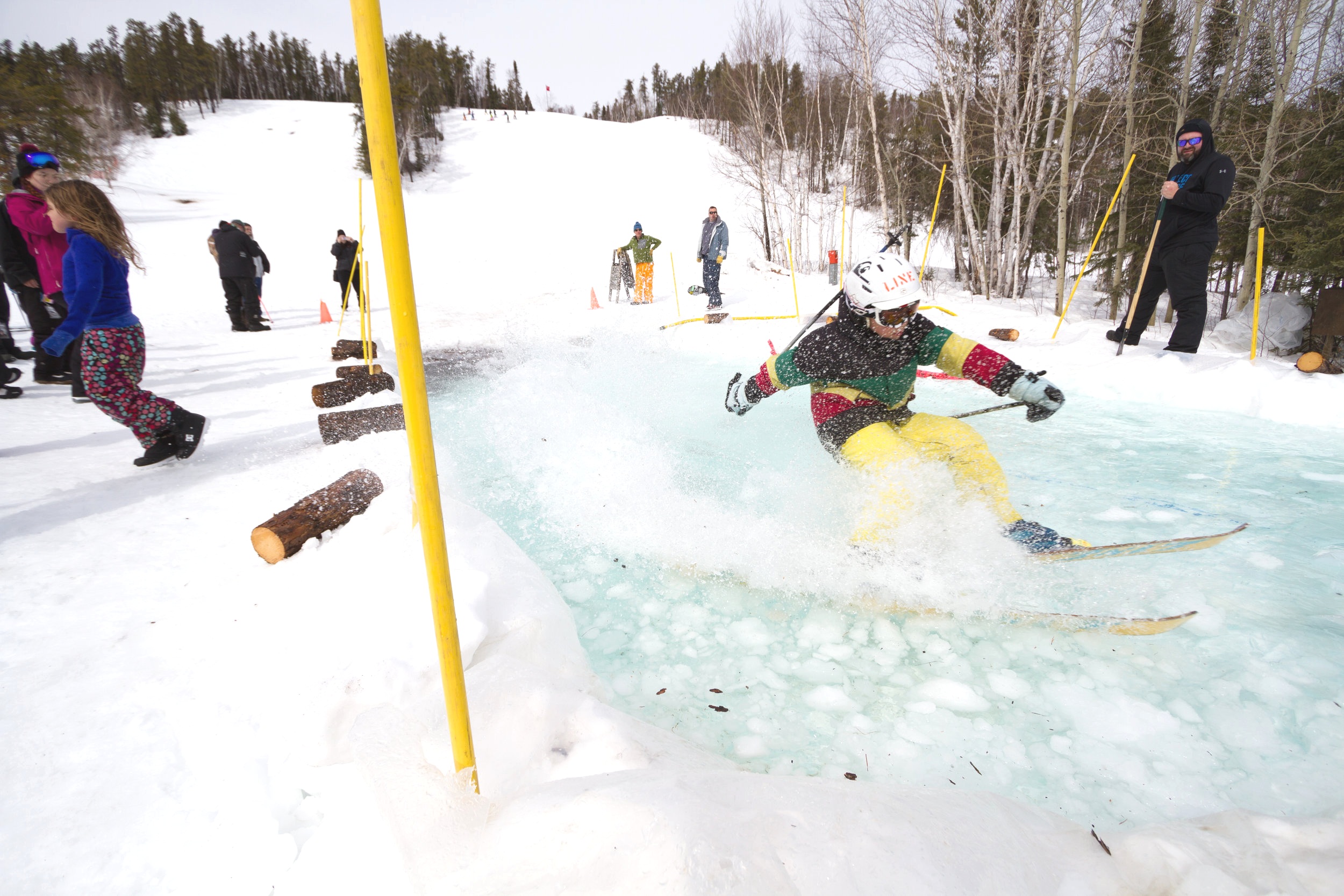 Winter sports scene at Mystery Mountain, Manitoba, Canada, prominently featuring a skier and a snowboarder in action, with a sports centre and a snowmobile faintly visible in the background.