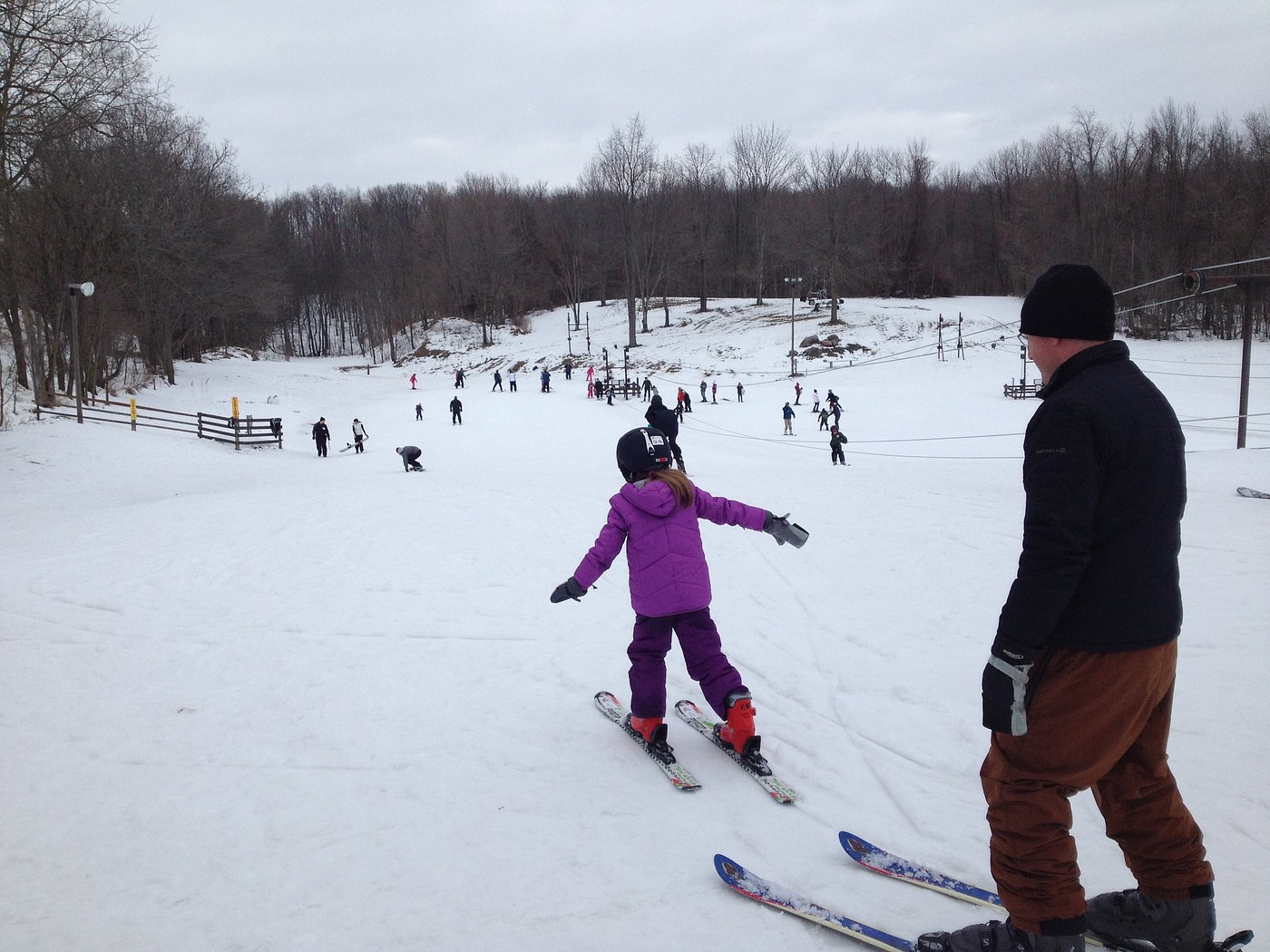 Winter sports scene at Swiss Valley, Michigan featuring a skier in the foreground. Families and individuals, including a learning child and a snowboarder, enjoy the snowy slopes.