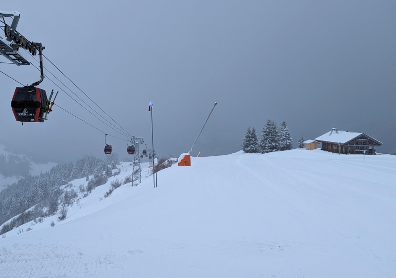 Skiers enjoying a sunny day at Domaine Skiable des Contamines Montjoie in France. A ski lift ascends the mountain, and a quaint challet sits in the distance.