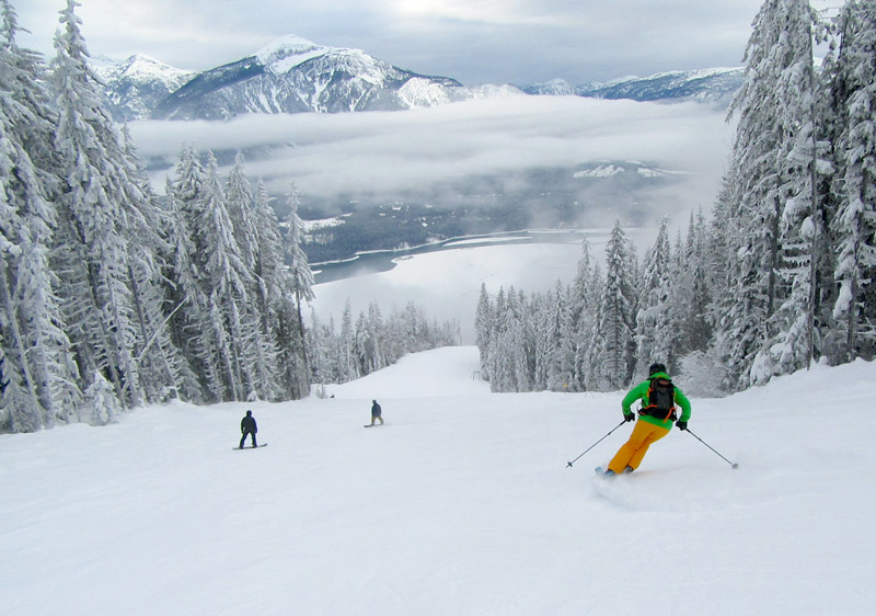 A skier descending a slope at the scenic Revelstoke Mountain Resort in British Columbia, Canada, surrounded by fellow skiers. A ski lift is faintly visible in the background.