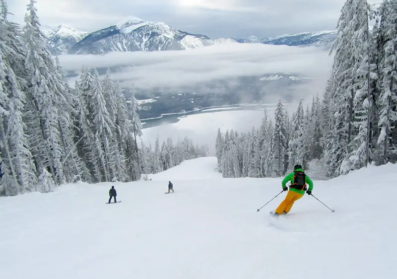 A skier descending a slope at the scenic Revelstoke Mountain Resort in British Columbia, Canada, surrounded by fellow skiers. A ski lift is faintly visible in the background.
