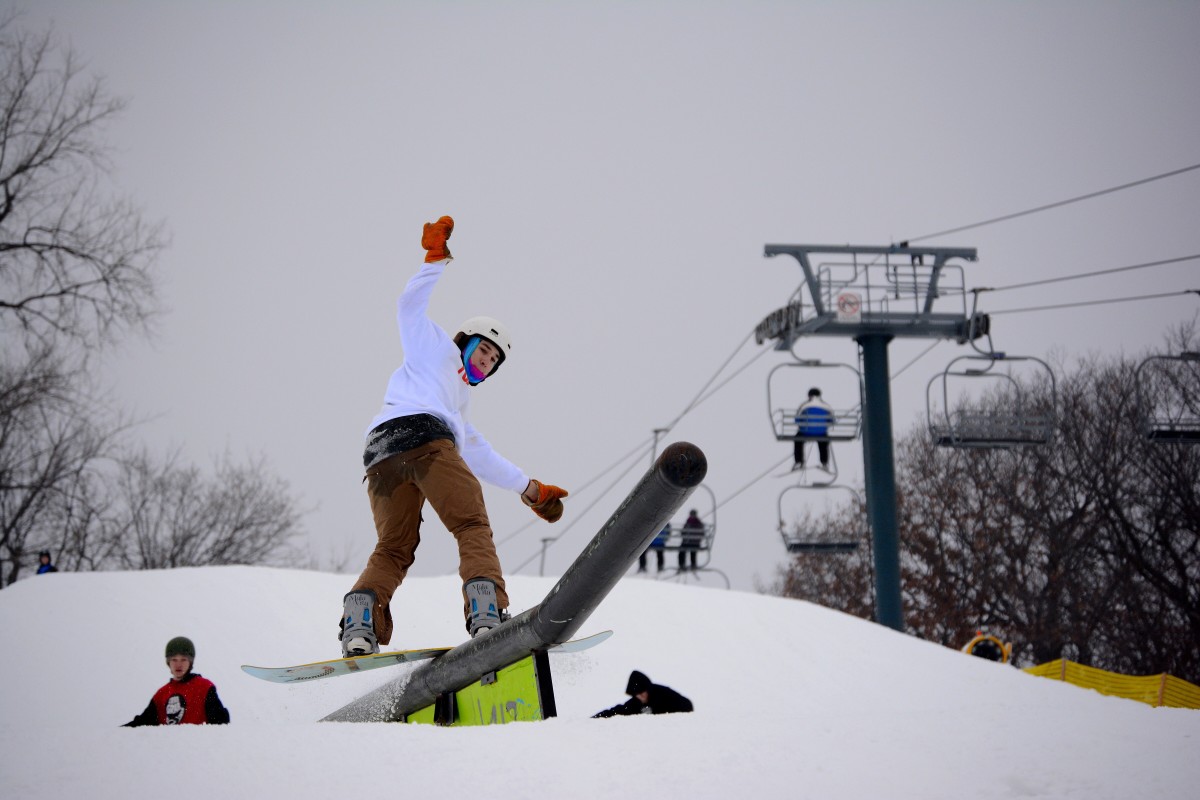 A snowboarder hitting the slopes at Buck Hill in Minnesota. He is mid-action with snow spraying around his board as he cuts through the white surface. The day is clear allowing a beautiful view of the hill.