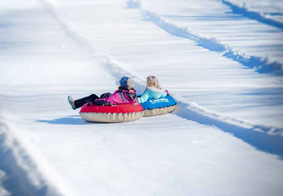 A winter sports scene at Yawgoo Valley Rhode Island featuring a snowmobile in the foreground. Visible in the background is a chalet and a family enjoying skiing.