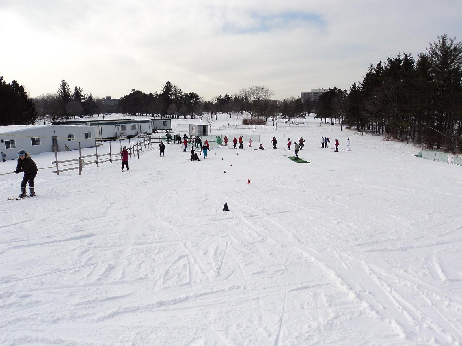 Winter sports scene at Earl Bales – North York, Ontario with a prominent winter sports centre, a distant view of a ski resort and chalet, and a skier huddled in warm gear, preparing for a downhill adventure.