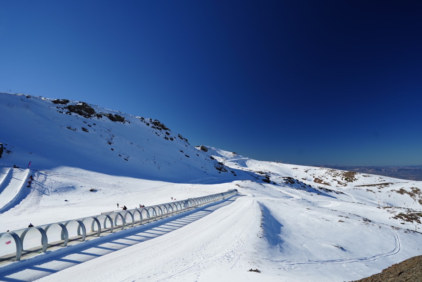 Winter scene in Sierra Nevada, Spain, showcasing gilt-edged mountains laden with snow, a bustling winter sports scene with a chalet and sports centre amid pristine, snowy landscape.