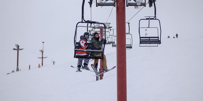 A ski lift ascends a snowy hill at The Rock Snowpark in Wisconsin USA carrying a skier and a snowboarder. Below another skier maneuvers the landscape while a challet peeks out in the background.