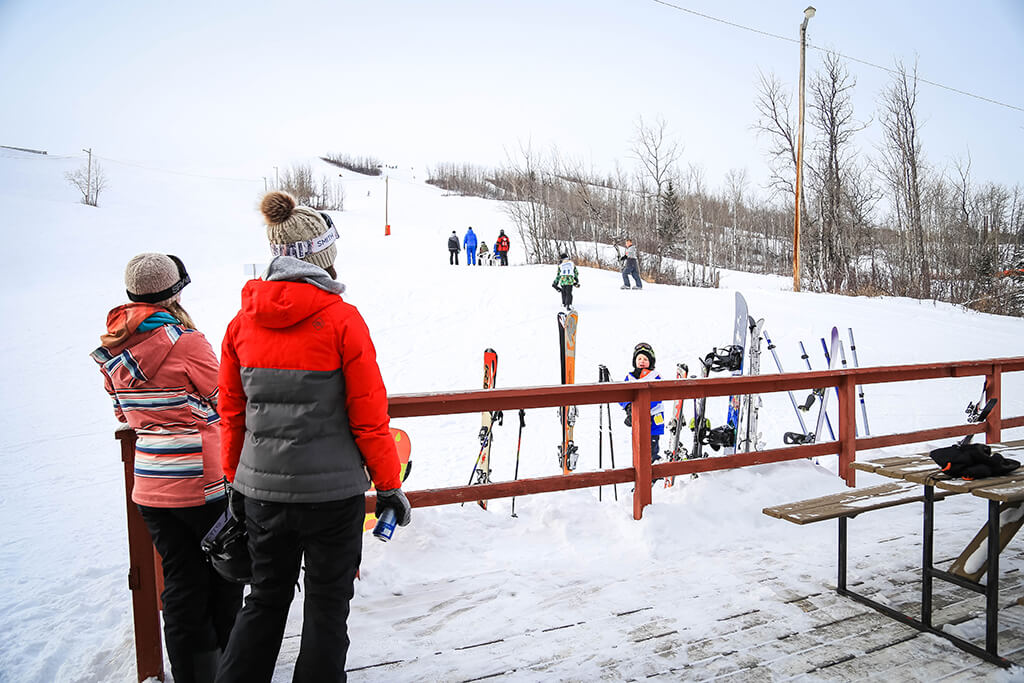 View of Fairview Ski Hill Alberta Canada during winter showcasing a skier in action surrounded by a lively winter sports scene. The ski resort and ski lift are also visible in the backdrop.