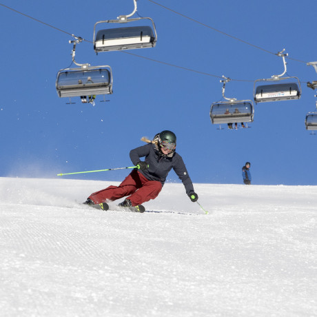 A skier gliding down a slope in Lachtal, Murtal, Styria, Austria, part of a bustling winter sports scene, with a ski lift and other snowboarders visible in the background of the ski resort.