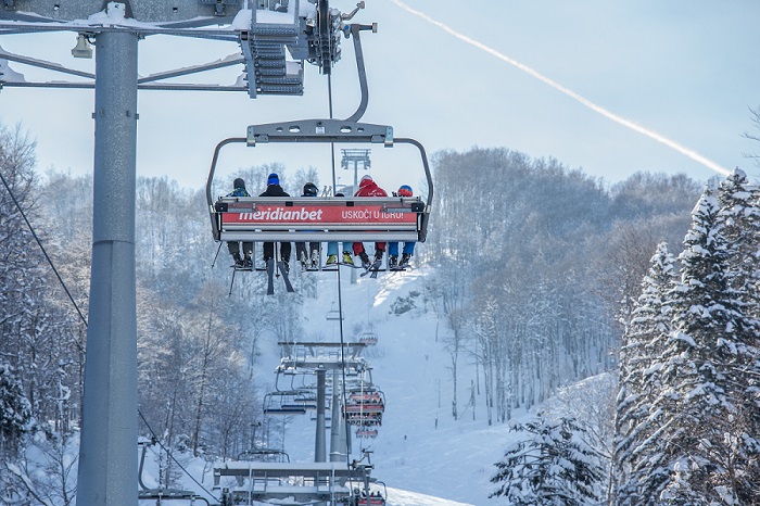 Image depicts a bustling ski resort in Kolašin, Montenegro with a ski lift operating against a stunning winter background, skiers descending down the snowy slopes, representing a vibrant winter sports scene.
