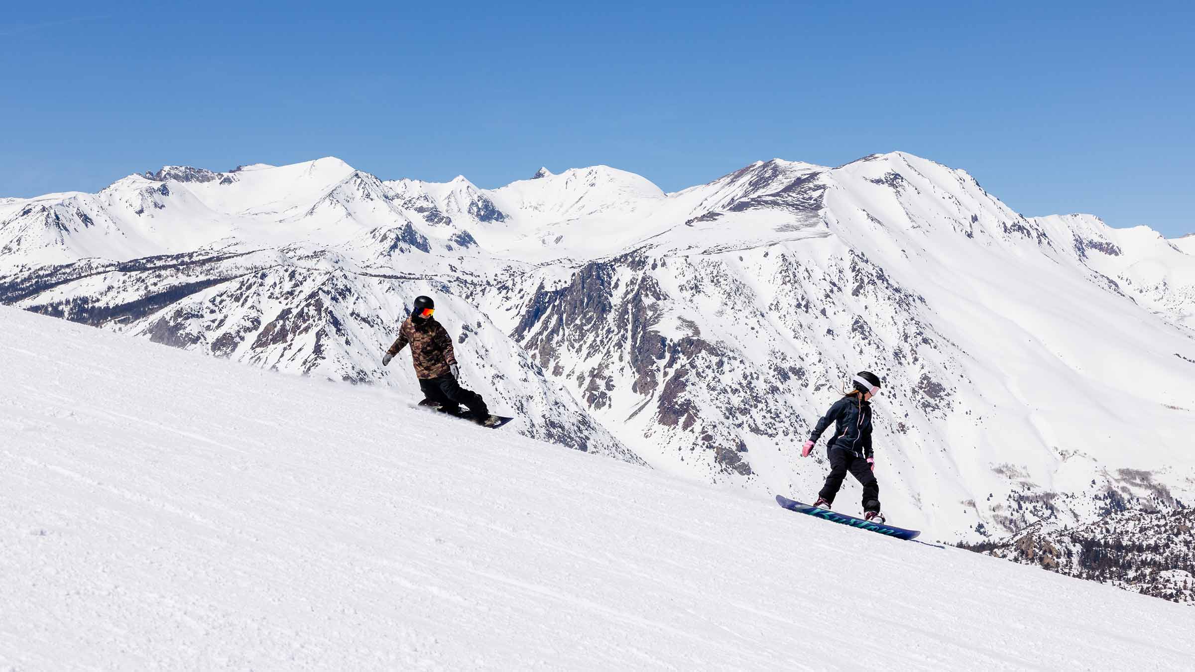 A snowboarder skillfully carving down a pristine hill at June Mountain in Mammoth Lakes, California. The white snow contrasts with the sunny, bright blue sky, creating a vibrant and adventurous scene.