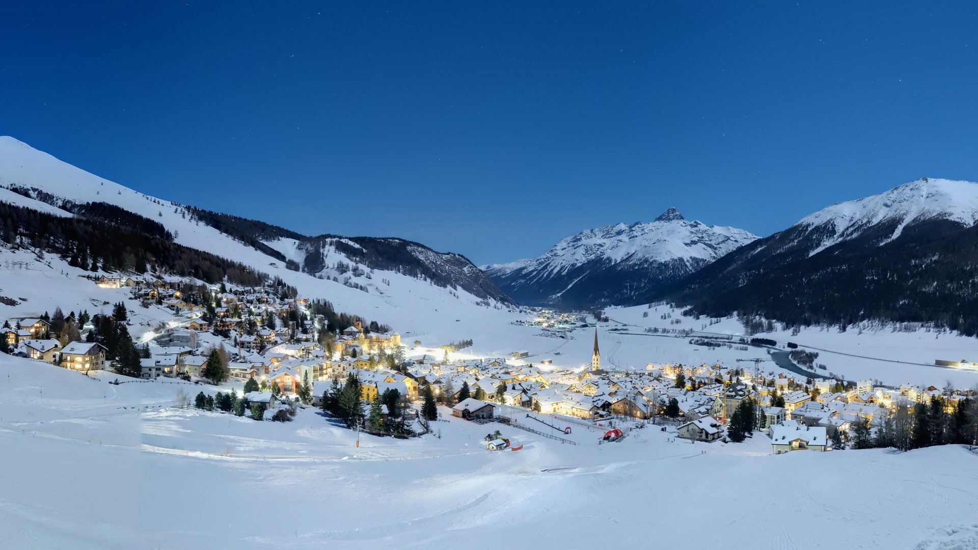 View of Zuoz – Pizzet | Albanas ski resort in Graubünden, Eastern Switzerland, showcasing a picturesque winter scene with bustling winter sports, surrounded by charming chalets.