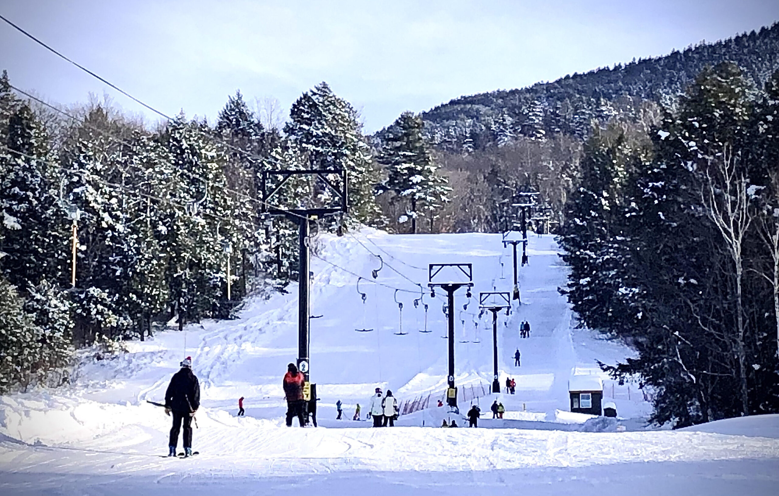 A bustling winter sports scene on Ascutney Mountain Vermont showcasing various skiers enjoying the picturesque ski trails with a ski lift in the distance and a ski resort faintly visible in the backdrop.