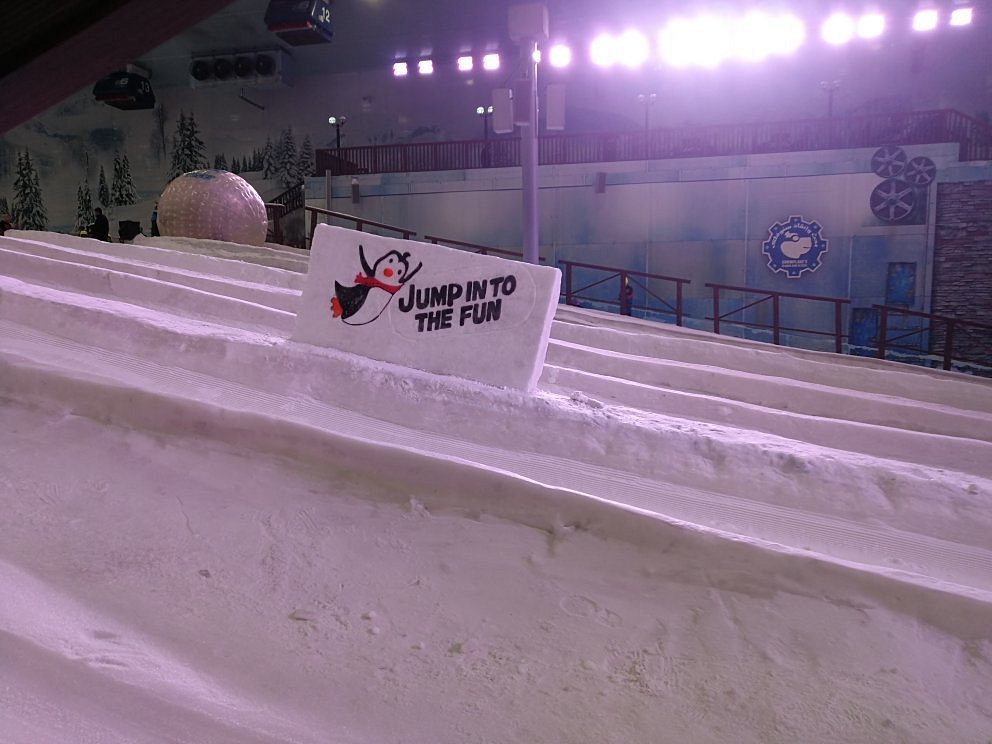 A skier glides past a chalet at Ski Egypt, a popular indoor winter sports centre. The image captures a vibrant winter sports scene with a snowmobile in the background.