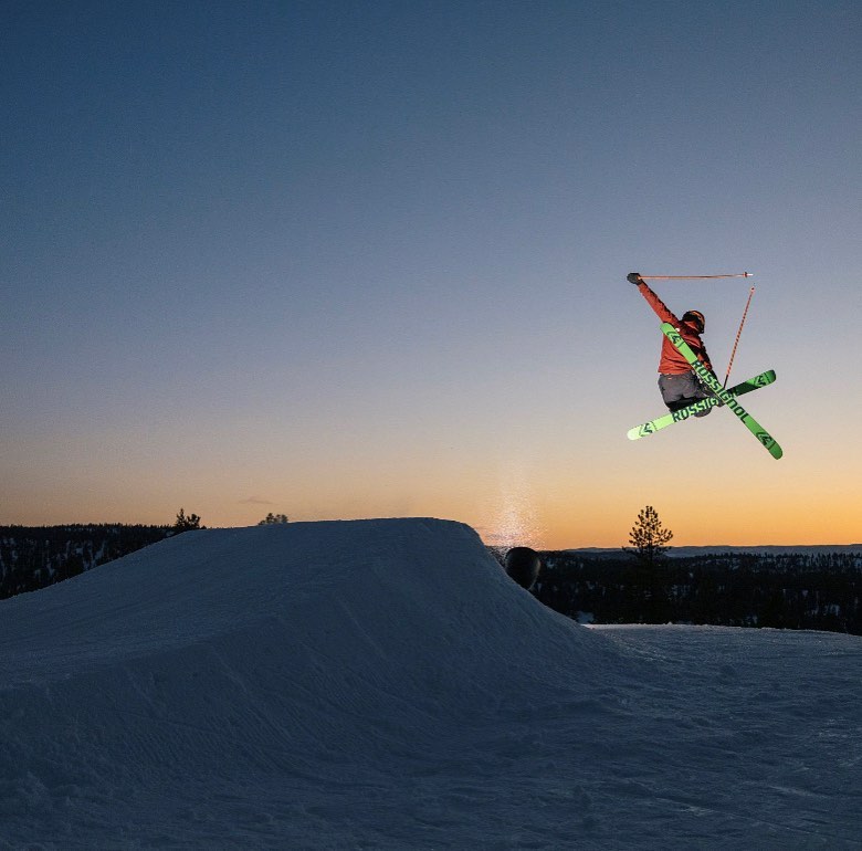 A skier enjoys an exhilarating run at Little Ski Hill Idaho carving through the snow amidst a bustling winter sports scene with a snowboarder in the distance and the resort's facilities subtly visible.