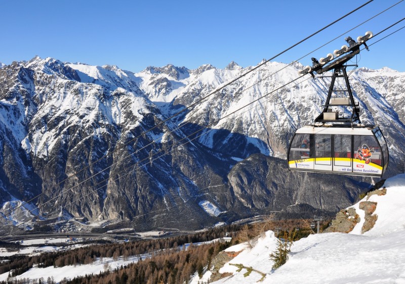 Winter image of Venet-Landeck, Tyrol, Austria showcasing a ski lift transporting visitors up to a ski resort. A charming chalet nestled in the snow sits nearby, as a skier enjoys the snowy landscape.