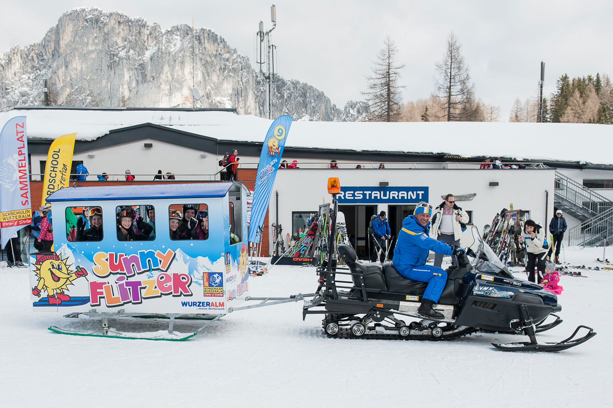 Picture captures a bustling winter sports scene at Wurzeralm – Spital am Pyhrn Austria with a ski lift various skiers and a snowmobile against the snowy backdrop of a popular ski resort.