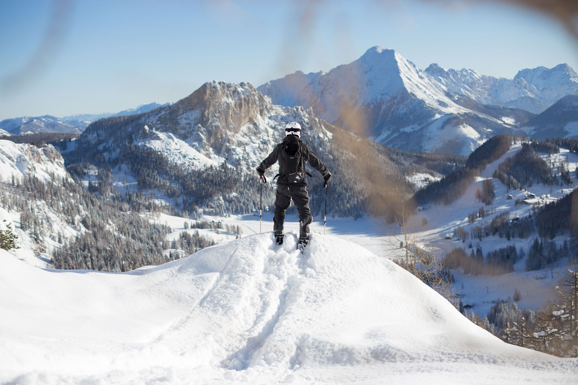 A winter sports scene at Wurzeralm – Spital am Pyhrn, Austria with a skier and snowboarder in action. A chalet nestled in the picturesque winter scenery in the background.