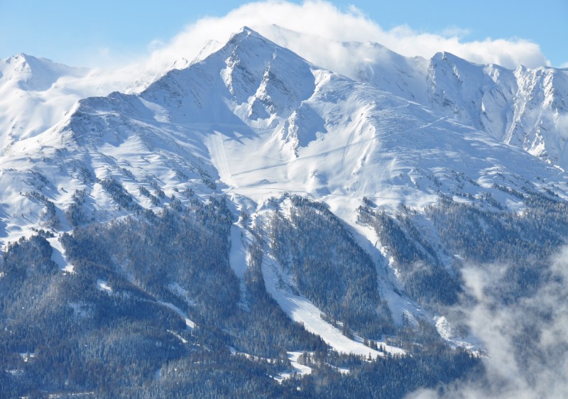 View of La Norma ski resort in Savoie Mont Blanc, France. Picture shows a towering mountain partially covered in snow with snow-swept slopes nearby. A cozy challet is also visible in the scene.