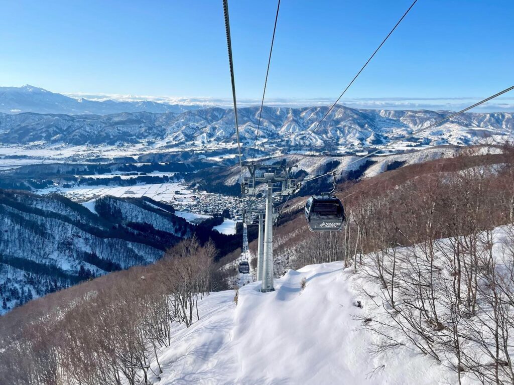 Winter scene at Nozawa Onsen in Japan featuring a bustling ski resort amidst a picturesque snowy landscape. Skiers are seen using a ski lift against a backdrop of pristine white, highlighting active winter sports.