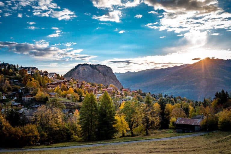 View of a picturesque chalet in Ovronnaz, Valais, Switzerland on a sunny day. The chalet sits against the backdrop of a snowy mountain, hinting at a nearby ski resort and winter sports activities.