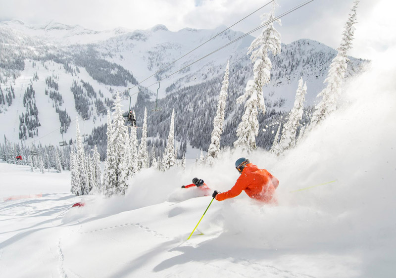 A skier going downhill at the Whitewater - Nelson ski resort in Kootenay Rockies, British Columbia, surrounded by snow and a ski lift visible in the distance.