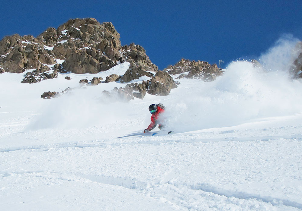 A skier swiftly gliding down a snowy slope in Las Leñas Argentina with a charming chalet and other winter sports enthusiasts visible in the background.