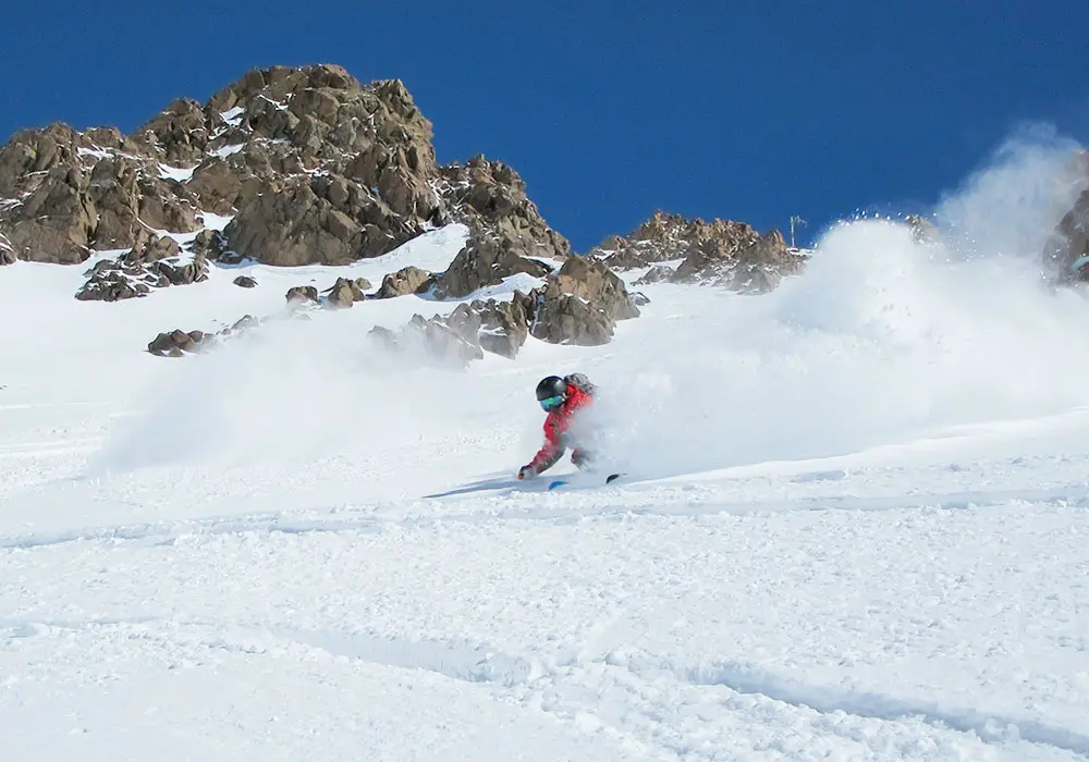 A skier swiftly gliding down a snowy slope in Las Leñas Argentina with a charming chalet and other winter sports enthusiasts visible in the background.