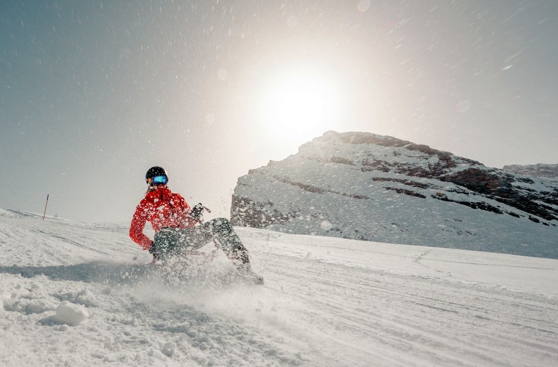Skier and snowboarder enjoying winter sports in the snowy landscape of Leukerbad, Switzerland, with chalets and snowmobiles nearby.