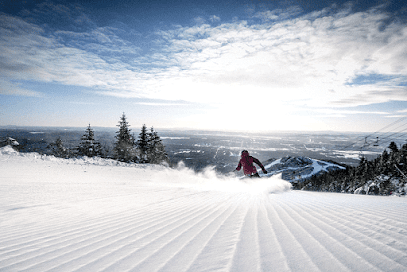 A skier gliding down a snowy mountainside at Mont-Orford in Quebec, Canada. The winter sports scene also features glimpses of a ski resort and a distant snowmobile.