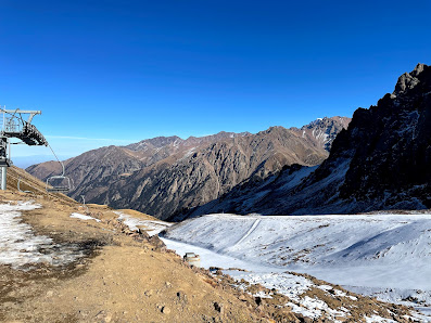 View of Shymbulak in Almaty, Kazakhstan depicting a picturesque mountain range. A chalet looms in the forefront with an active winter sports scene around it, enhancing the stunning winter scenery.