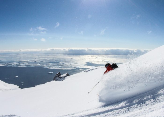 A skier skillfully gliding down a snowy mountainside on Disko Island in Greenland. The scene highlights the serene winter sports environment along with glimpses of a ski resort and a chalet in the distance.