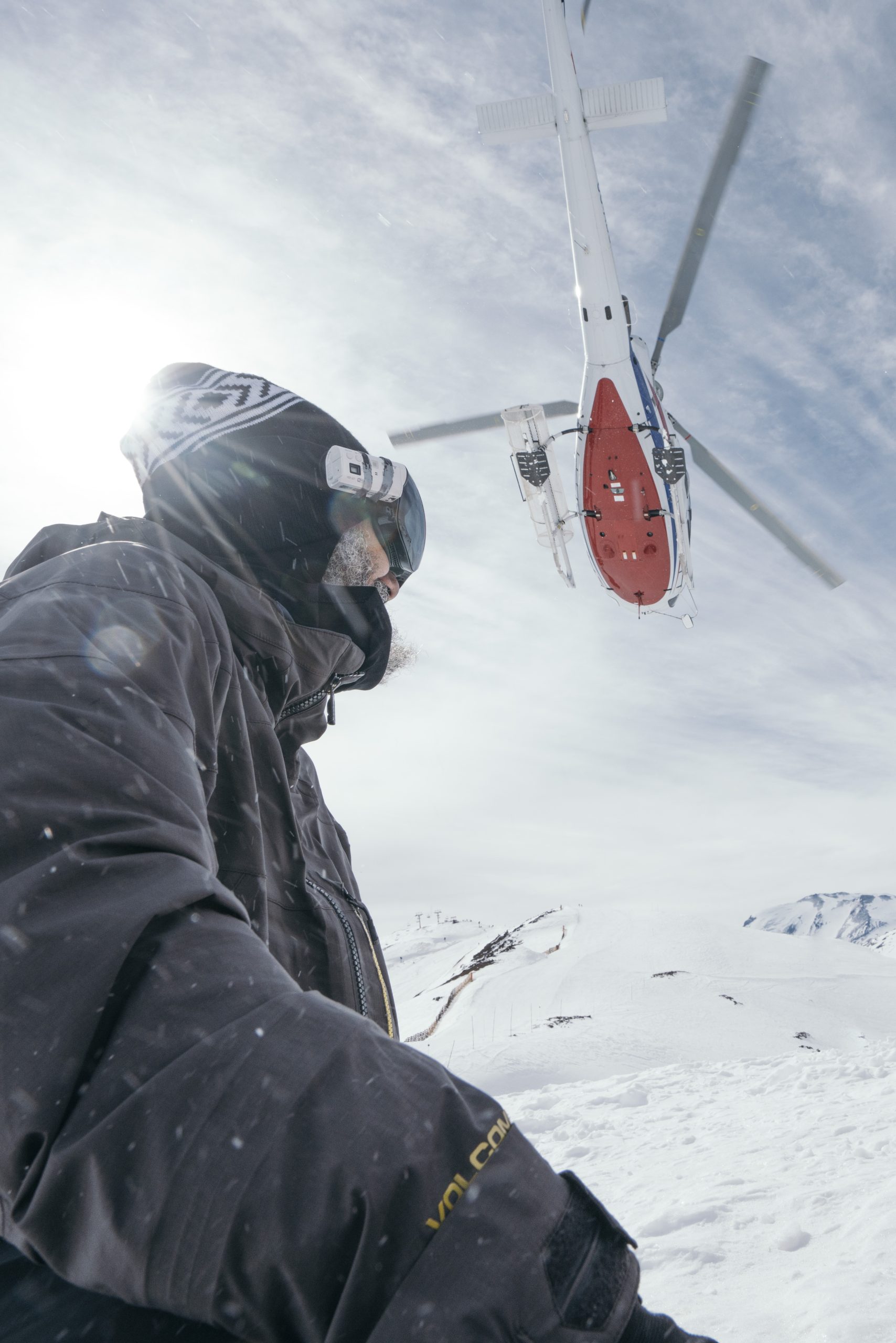 A skier gliding down the snowy slopes of Valle Nevado in Santiago, Chile with a snowboarder in distance. A ski lift trails up the mountainside, leading to a cozy chalet.