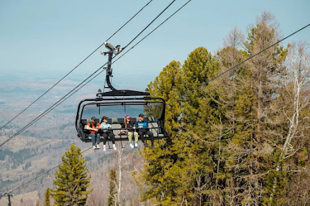 A winter scene at Manzherok in Ozernoe Siberia featuring a ski lift traversing the snowy terrain with a skier making their way down the slopes. The setting suggests it's a part of a ski resort.