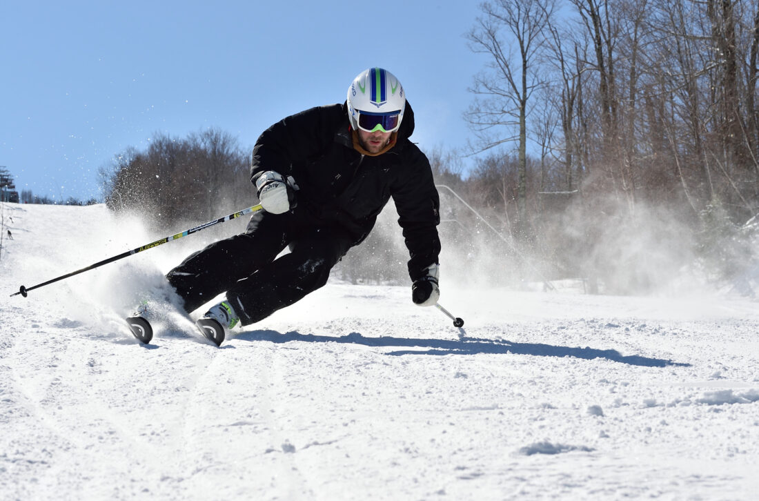 A skier enjoys a winter sports scene at Owl's Head in Mansonville, Quebec, with a glimpse of a snowboarder in the snow-covered landscape. A cozy challet completes the picturesque setting.