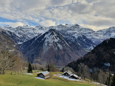 A scenic view at San Bernardino Pian Cales, Eastern Switzerland featuring a cozy chalet surrounded by a captivating winter scenery. The backdrop is set against a majestic mountain range, hinting at a nearby ski resort.
