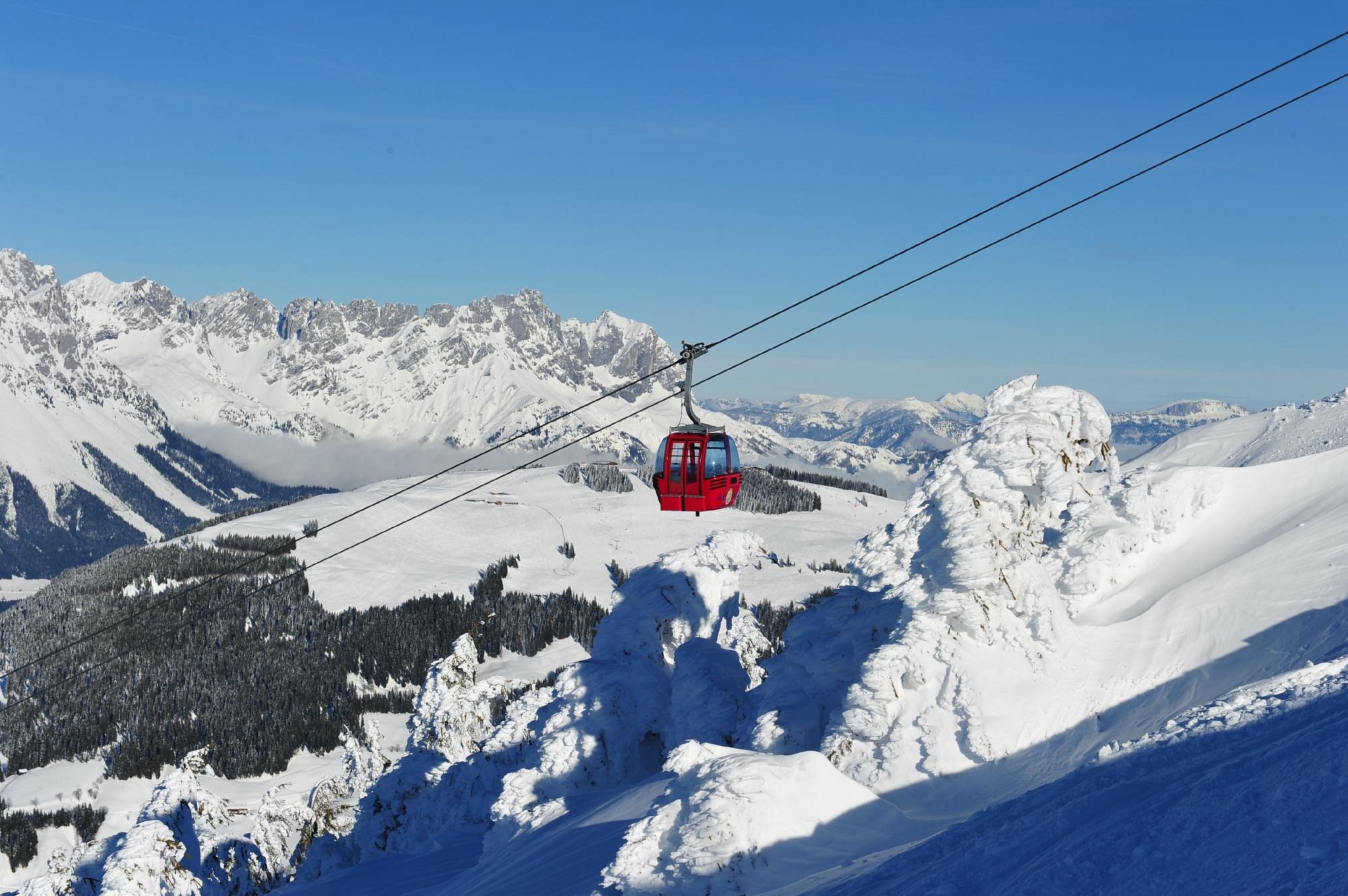 Ski lift in operation at the SkiWelt -Brixental resort in Tyrol, Austria, amidst a bustling winter sports scene. A chalet can be seen in the background and a skier is in motion.