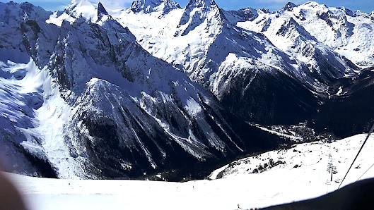 View of Dombai in Caucasus, featuring a large mountain with a snowy peak and a challet nestled at the base. It's a winter sports scene at a ski resort, with the landscape blanketed in snow.