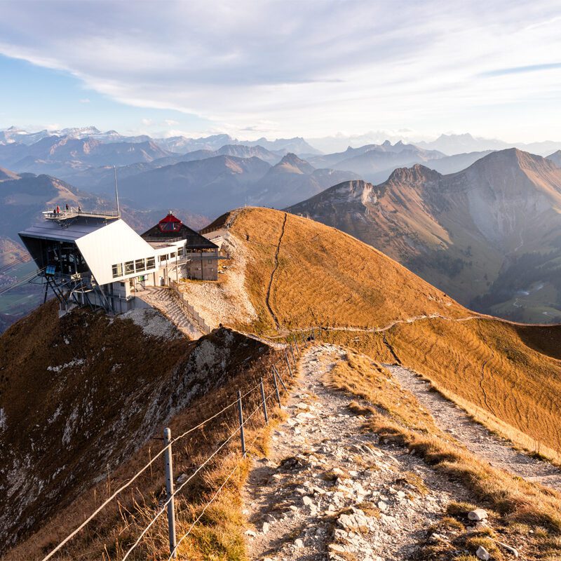 A picturesque swiss scene at Moléson in Fribourg featuring a cozy mountain hut and chalet nestled amidst a prominent mountain range. The landscape indicates popular winter sports and ski resort activities.
