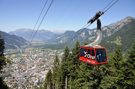 A ski lift ascends a snowy Swiss mountain in Chur Graubünden at the Brambrüesch - Chur Bergbahnen ski resort with a charming chalet nearby and a lone skier hitting the slopes.