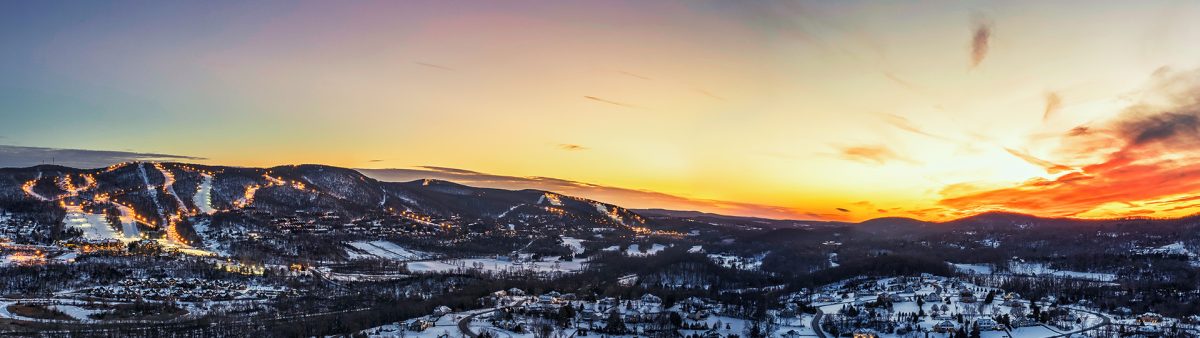 Snowy scene at Mountain Creek – Vernon, New Jersey featuring a bustling winter sports scene with skiers and a chalet nearby. A ski lift transports visitors overhead, portraying an active ski resort landscape.