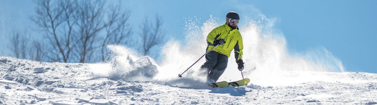 A skier is skillfully navigating down the slopes at the Mountain Creek – Vernon ski resort in Vernon Township New Jersey surrounded by a typical winter sports scene with hints of a snowboarder and snowmobile in the background.