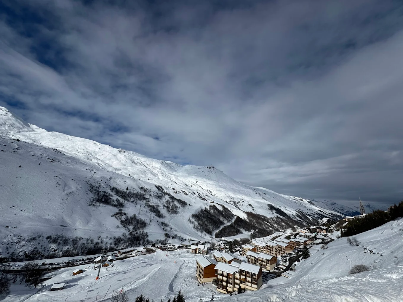 A skier enjoying winter sports at Les 3 Vallées in France, surrounded by magnificent snowy peaks. Nearby is a family skiing and a distant glimpse of a snowboarder. A ski lift can also be spotted in the scene.