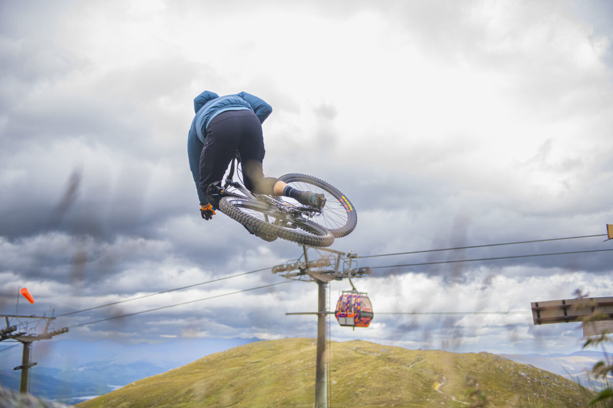 A mountain biker traversing the rugged terrain of Nevis Range in the Highlands, Scotland, with a ski lift and challet in the background. A distantly spotted skier adds to the winter sports scene.