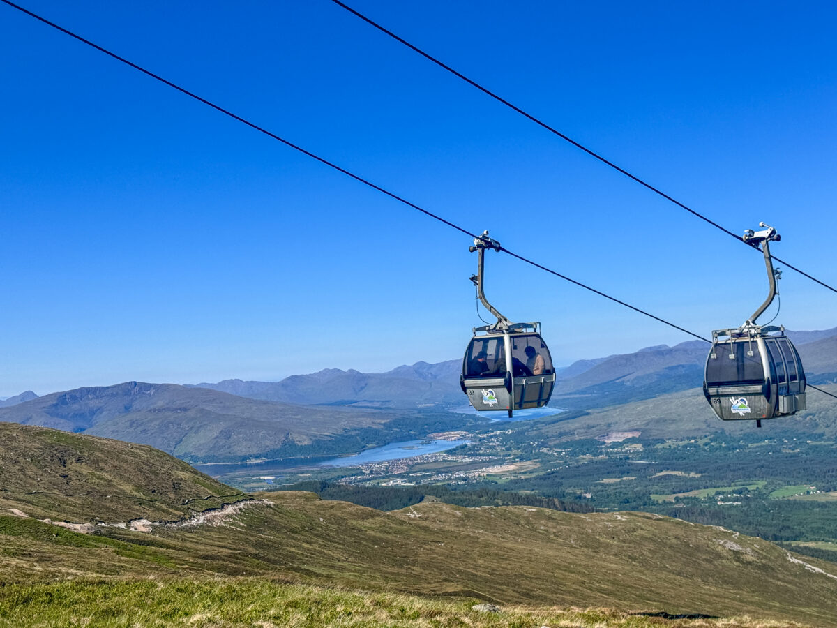 Ski lift at the Nevis Range in the Scottish Highlands, with a chalet and a buzzing ski resort in the backdrop on a bright sunny day.