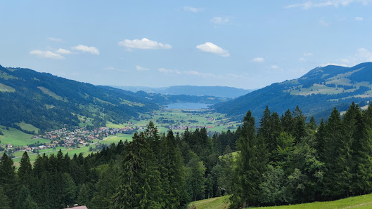A charming chalet overlooks a serene lake on a sunny day in Hündle Thalkirchdorf Swabia Germany. The view is completed with a striking mountain backdrop and hints of a ski resort.