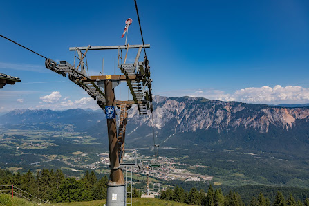 View of a ski lift ascending up a mountain at Arnoldstein Dreiländereck in Austria indicating the presence of a ski resort. Skiers and winter sports can also be seen in the scene.